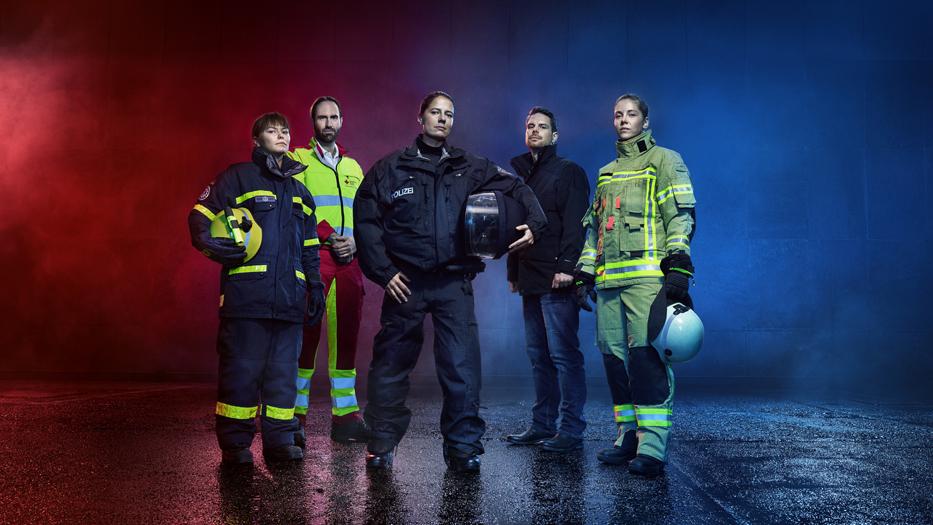 Vor einem blau-roten Hintergrund stehen 5 Rettungskräfte: ein Kriminalbeamter,...